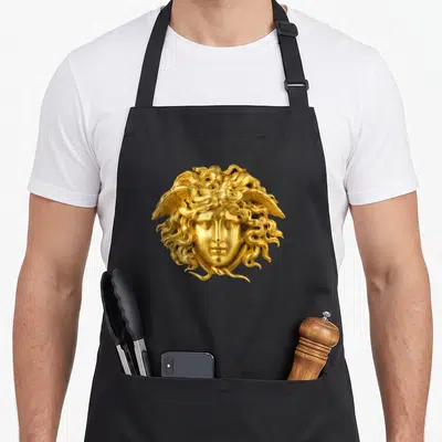 Snake hair apron - Golden sculpted Medusa head with snake hair and wings centered on grey background — shown on a black cotton canvas apron against a white background