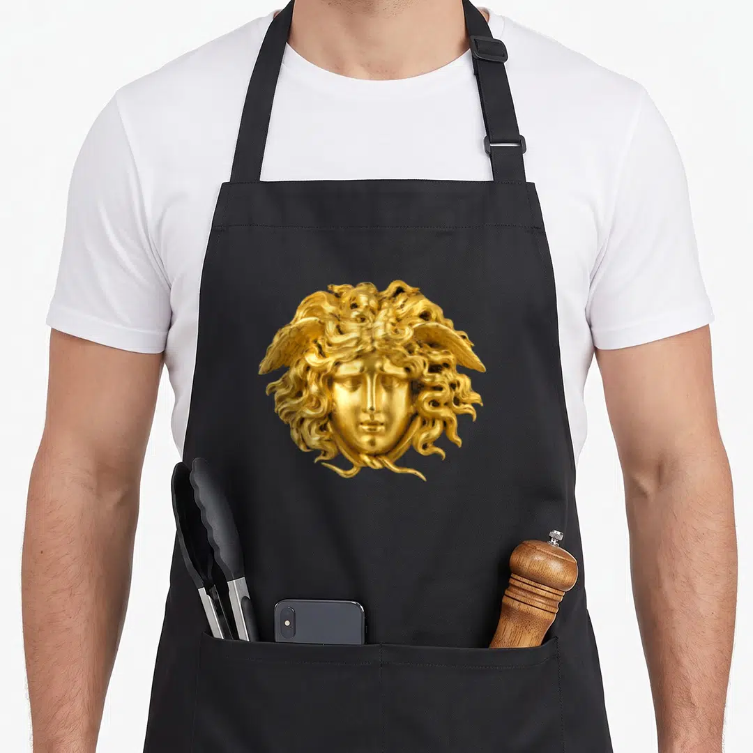 Snake hair apron - Golden sculpted Medusa head with snake hair and wings centered on grey background — shown on a black cotton canvas apron against a white background