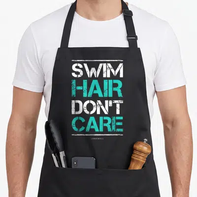 Swim hair apron - Distressed white and teal stacked text reading SWIM HAIR DON'T CARE framed by horizontal lines — shown on a black cotton canvas apron against a white background