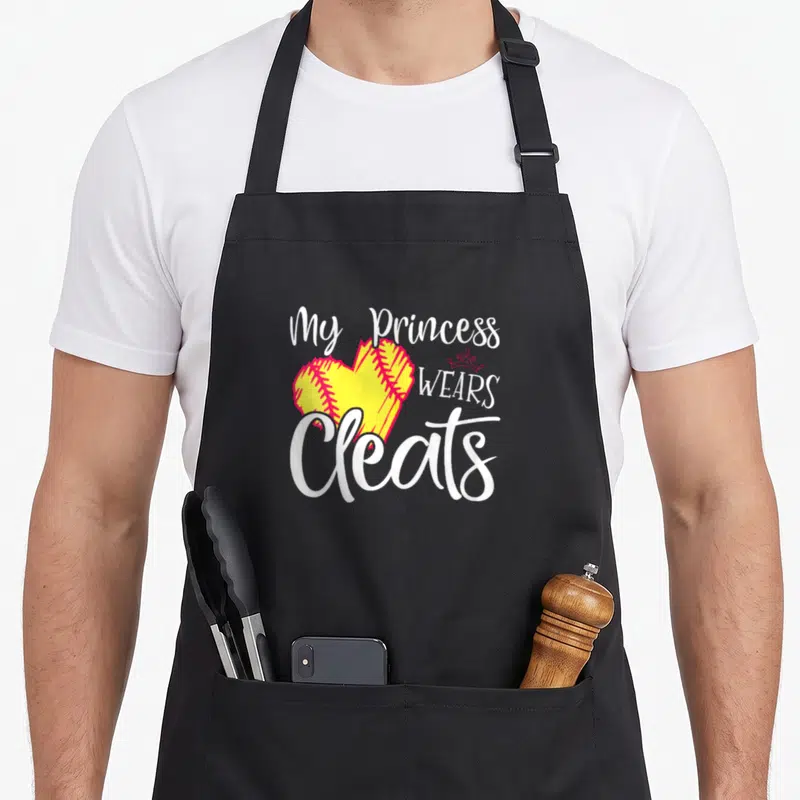 Softball princess apron - Yellow softball heart with red stitching and white text reading My Princess Wears Cleats — shown on a black cotton canvas apron against a white background
