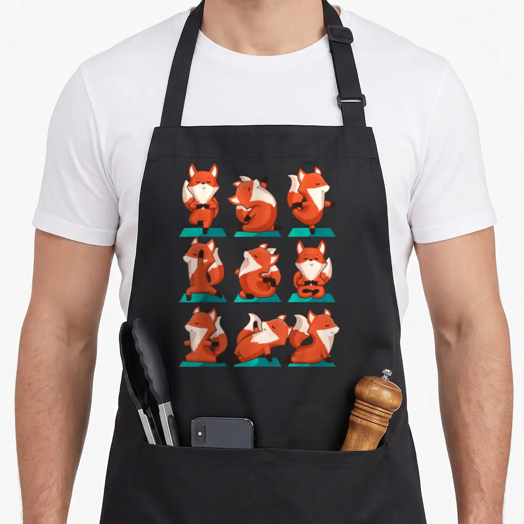 Fox grill instructor apron - Nine cartoon foxes in various yoga poses on teal mats arranged in a grid — shown on a black cotton canvas apron against a white background