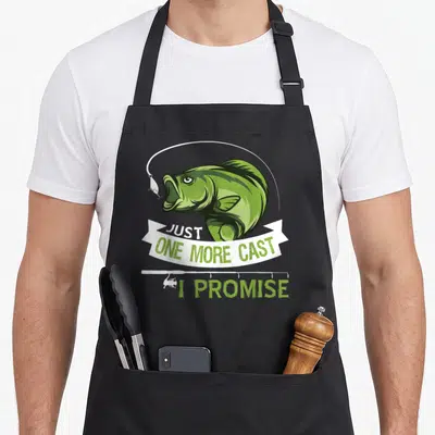 Cast fishing aprons - Green cartoon fish head over white banner reading JUST ONE MORE CAST I PROMISE with fishing rod — shown on a black cotton canvas apron against a white background