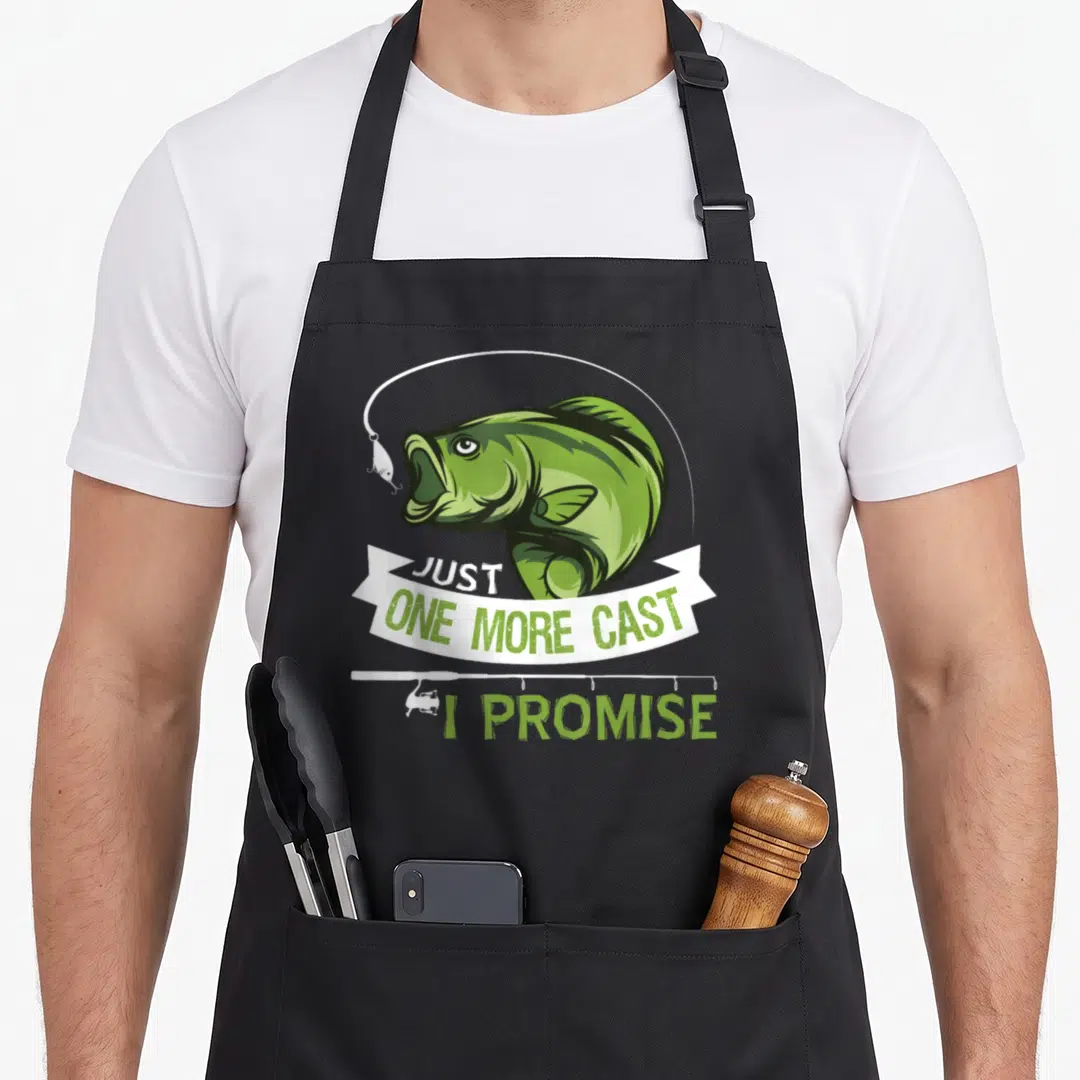 Cast fishing aprons - Green cartoon fish head over white banner reading JUST ONE MORE CAST I PROMISE with fishing rod — shown on a black cotton canvas apron against a white background