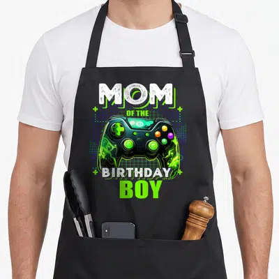Bday video game apron - MOM OF THE BIRTHDAY BOY text surrounding a neon green and black game controller on a blue grid — shown on a black cotton canvas apron against a white background