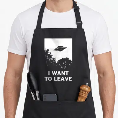 Believer alien ufo apron - Grey UFO and tree silhouettes in white frame above I WANT TO LEAVE text — shown on a black cotton canvas apron against a white background