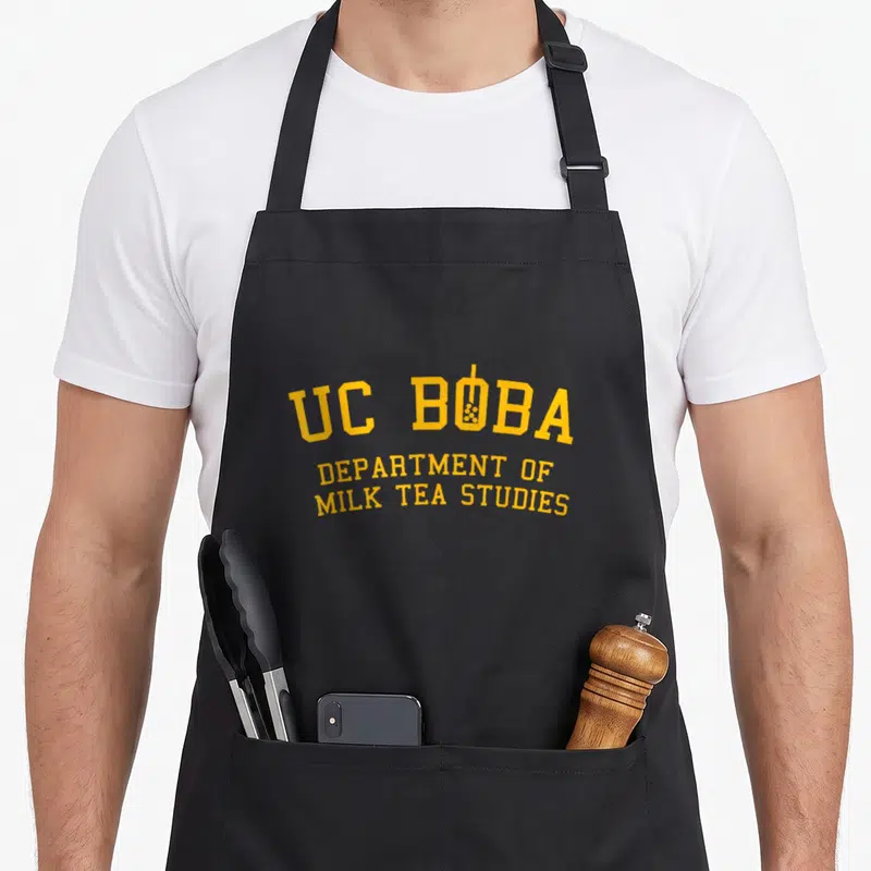 Milk asian apron - Yellow UC BOBA text with boba cup icon replacing the letter O — shown on a black cotton canvas apron against a white background
