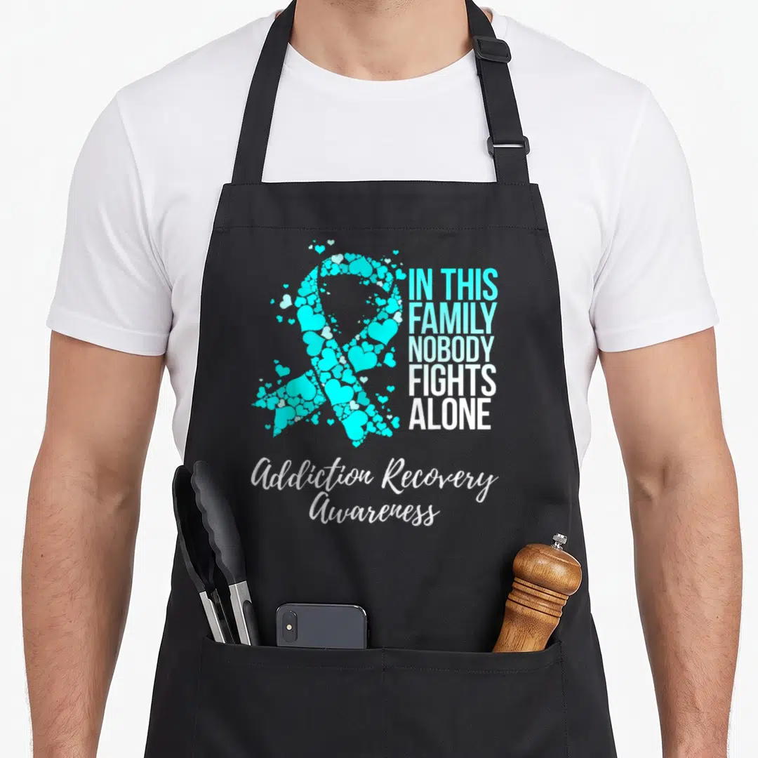 Addiction family aprons - Light blue heart ribbon and IN THIS FAMILY NOBODY FIGHTS ALONE text with script below — shown on a black cotton canvas apron against a white background