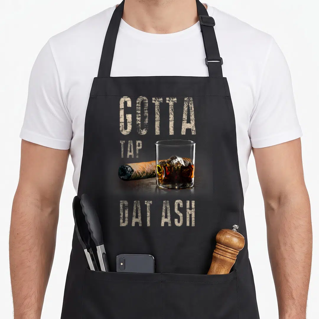 Cigar smoking apron - Beige text Gotta Tap Dat Ash above whiskey glass and brown cigar photo — shown on a black cotton canvas apron against a white background