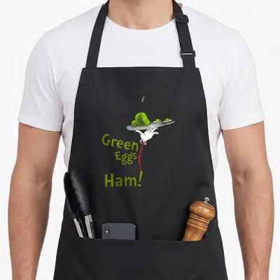 Title egg apron - Cartoon hand holding plate with green food and stacked text connected by red line — shown on a black cotton canvas apron against a white background