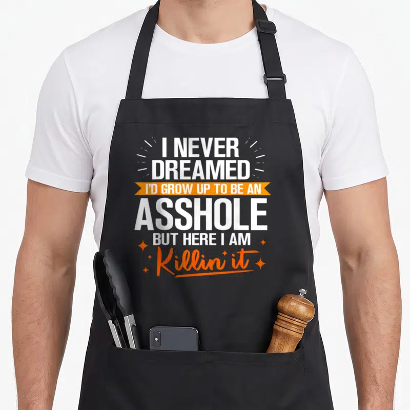 Funny grow never dreamed apron - Stacked white text on orange ribbon flanked by four sparkles — shown on a black cotton canvas apron against a white background