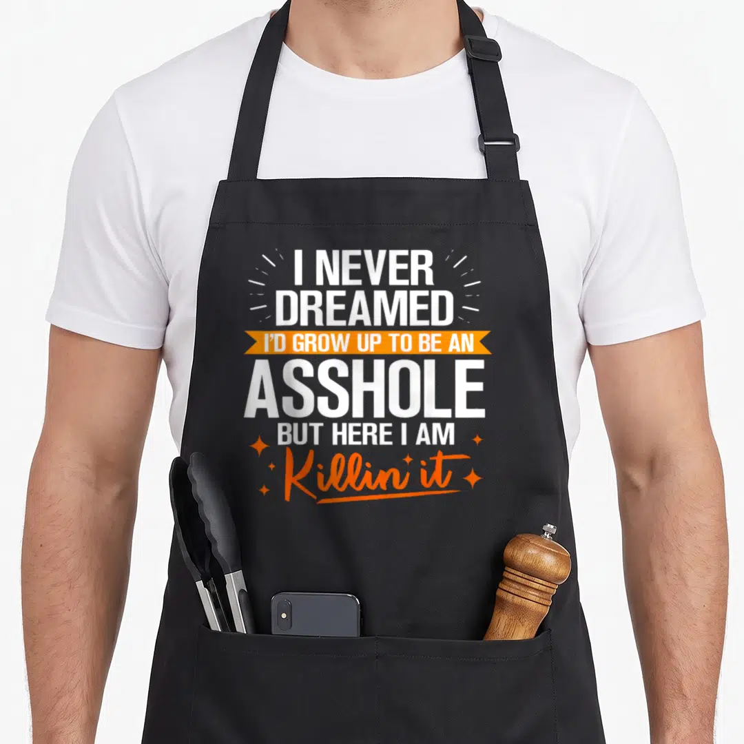 Funny grow never dreamed apron - Stacked white text on orange ribbon flanked by four sparkles — shown on a black cotton canvas apron against a white background