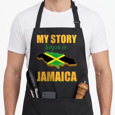Jamaican story of the apron - Map of Jamaica in yellow green and black with MY STORY began in JAMAICA text — shown on a black cotton canvas apron against a white background
