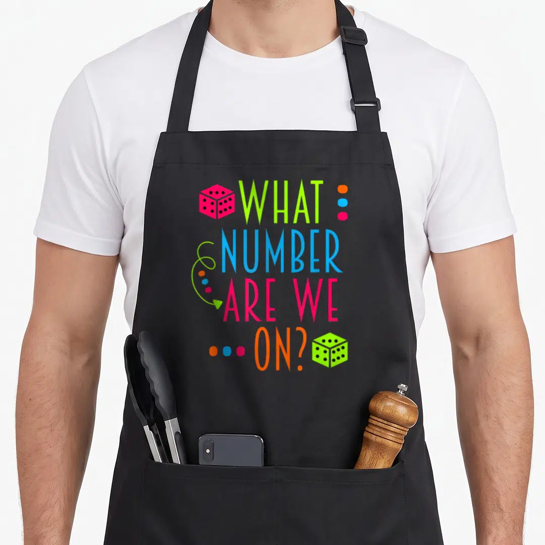 Number funny babe apron - Stacked colorful text what number are we on with pink and green dice and curly arrow — shown on a black cotton canvas apron against a white background