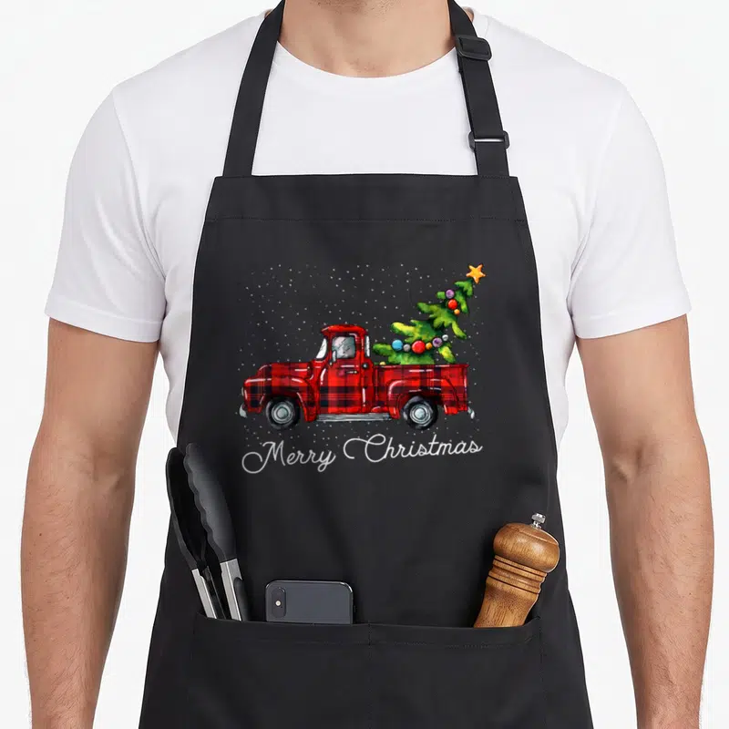 Truck buffalo plaid christmas apron - Red plaid truck carrying green Christmas tree with white Merry Christmas script — shown on a black cotton canvas apron against a white background