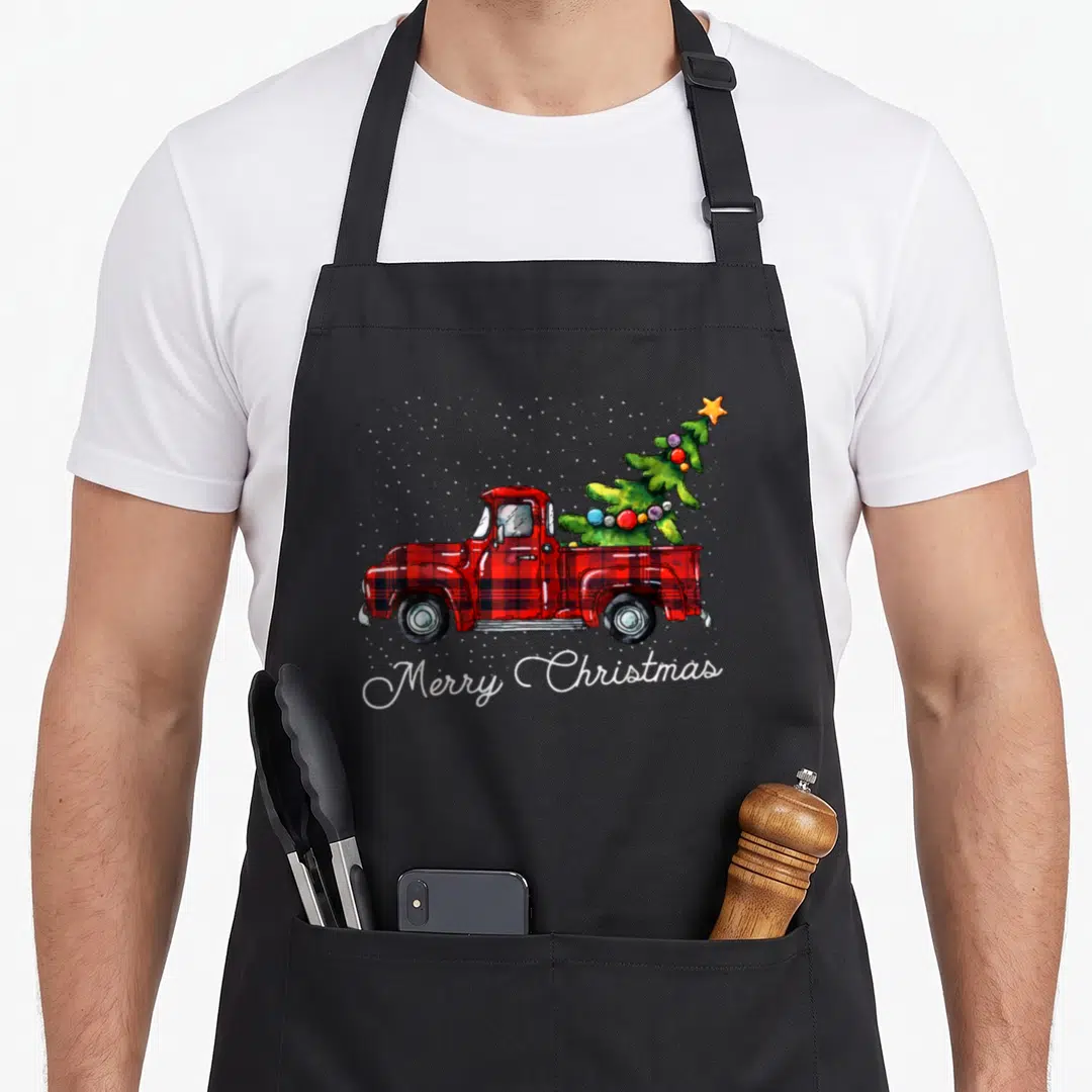 Truck buffalo plaid christmas apron - Red plaid truck carrying green Christmas tree with white Merry Christmas script — shown on a black cotton canvas apron against a white background
