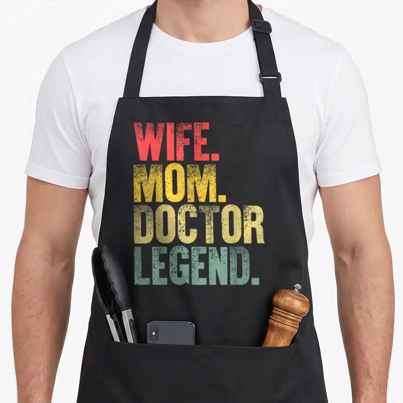 Doctor mother's apron - Stacked WIFE. MOM. DOCTOR. LEGEND. text in red yellow and teal distressed fonts — shown on a black cotton canvas apron against a white background