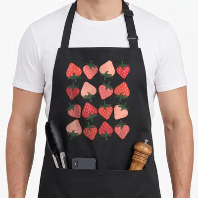 Pattern strawberry apron - Sixteen red pink and green cartoon strawberries in a 4x4 grid pattern — shown on a black cotton canvas apron against a white background