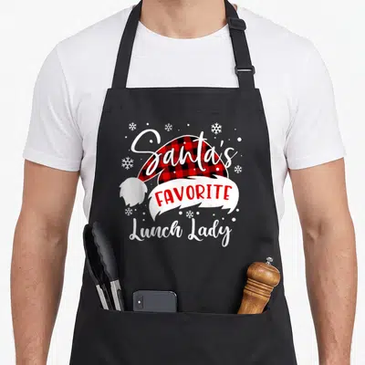 Santas lunch lady apron - Santa hat with red black plaid and Santa's Favorite Lunch Lady text surrounded by snowflakes — shown on a black cotton canvas apron against a white background
