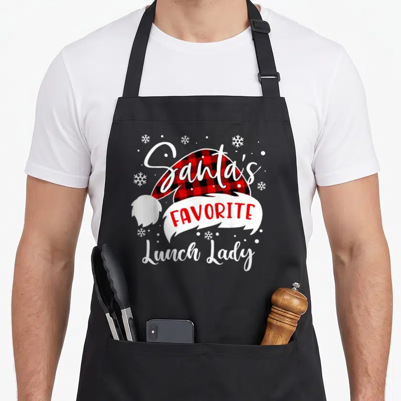 Santas lunch lady apron - Santa hat with red black plaid and Santa's Favorite Lunch Lady text surrounded by snowflakes — shown on a black cotton canvas apron against a white background