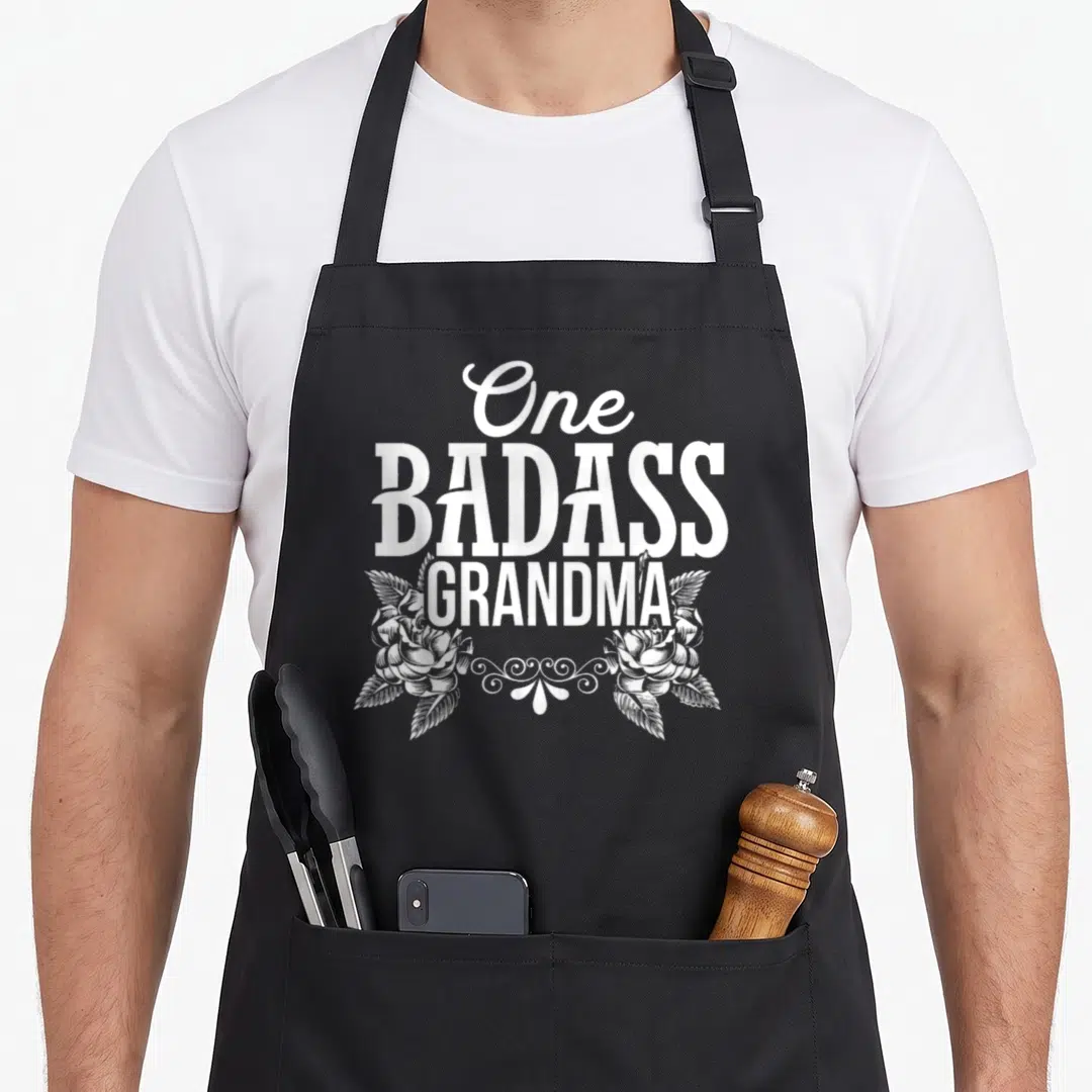 Grandma badass apron - White stacked text One BADASS GRANDMA above two roses and swirl ornament — shown on a black cotton canvas apron against a white background