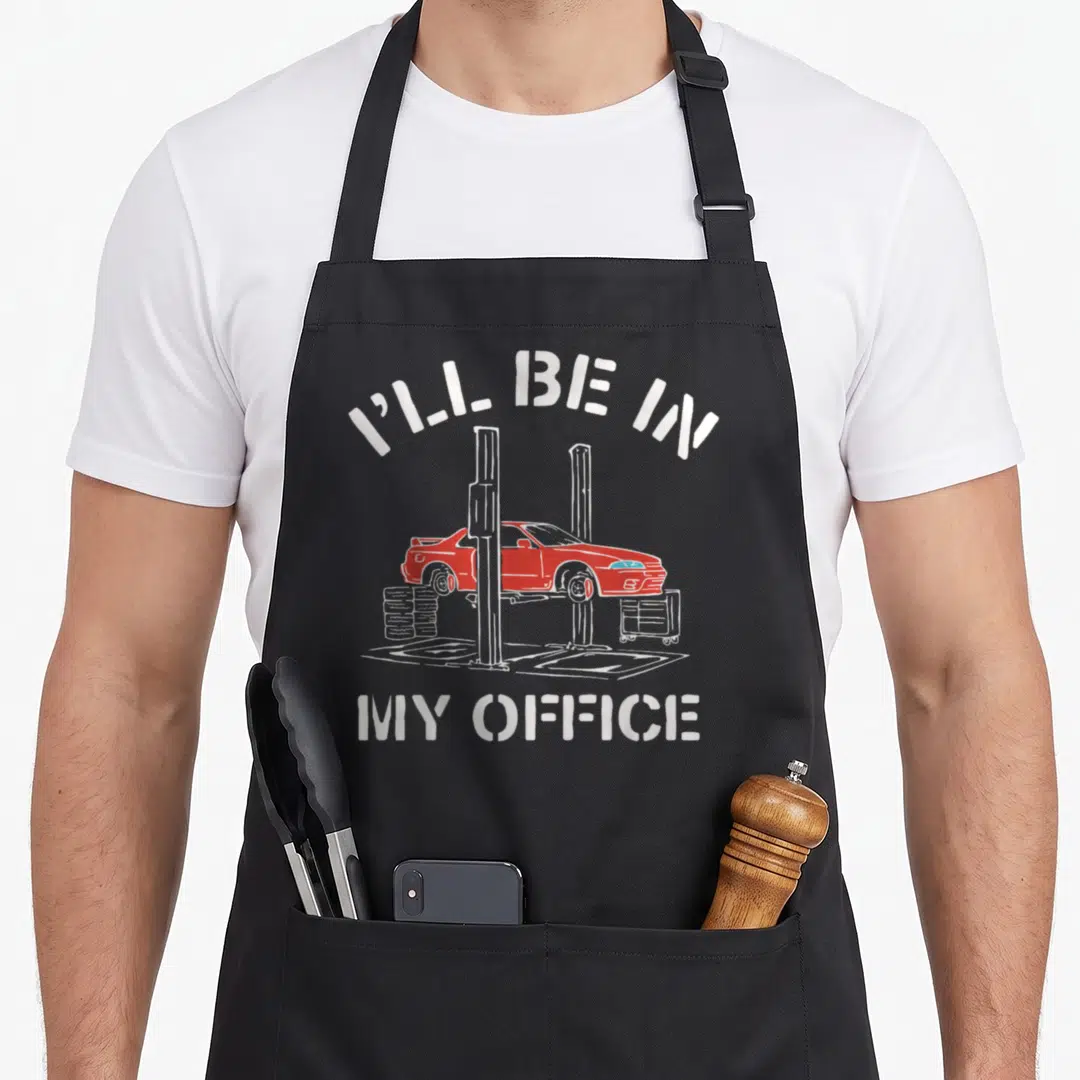 Ill auto mechanic apron - Red car on white lift with tires and toolbox under arched text reading I'll be in my office — shown on a black cotton canvas apron against a white background