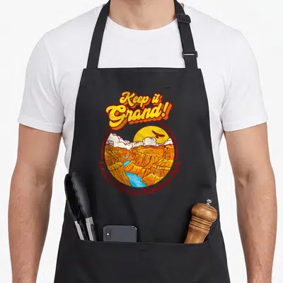 Grand desert camping apron - Retro Grand Canyon circle art with Keep it Grand text in orange yellow red blue — shown on a black cotton canvas apron against a white background
