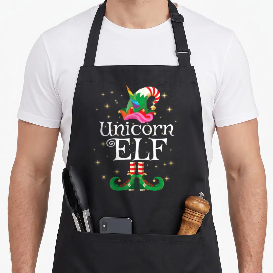 Unicorn christmas elf apron - Green elf hat with rainbow horn, white text Unicorn ELF, and curled green shoes with stars — shown on a black cotton canvas apron against a white background