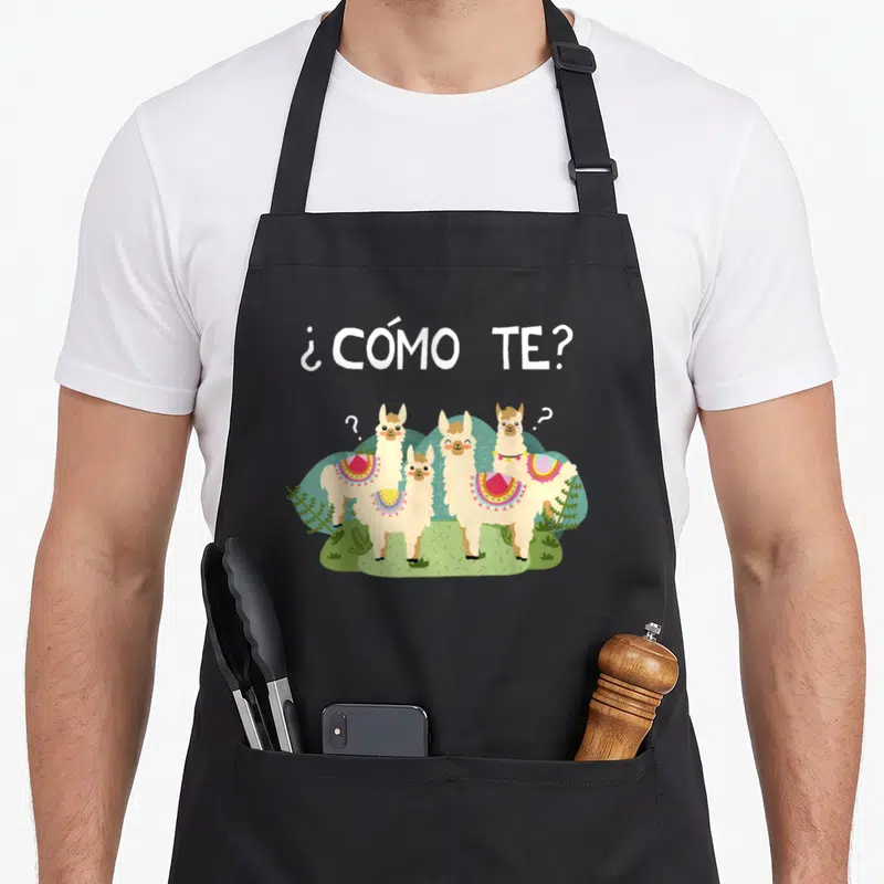 Llama spanish aprons - ¿CÓMO TE? text above four cartoon llamas on grass — shown on a black cotton canvas apron against a white background