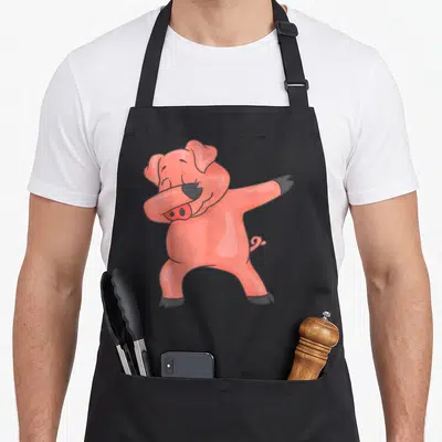 Dancing farmers apron - Pink cartoon pig dabbing pose with black outlines and hooves — shown on a black cotton canvas apron against a white background