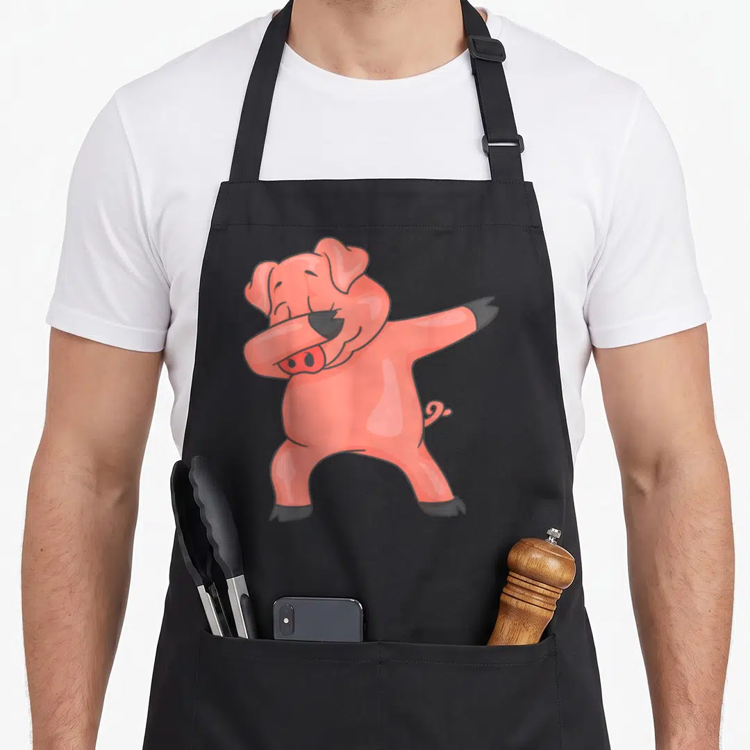 Dancing farmers apron - Pink cartoon pig dabbing pose with black outlines and hooves — shown on a black cotton canvas apron against a white background