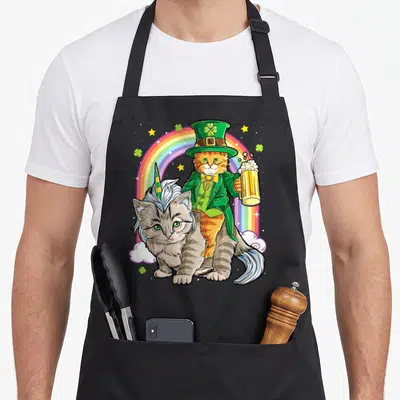 Riding st patrick day aprons - Orange cat in green suit riding grey horned cat under rainbow with shamrocks — shown on a black cotton canvas apron against a white background