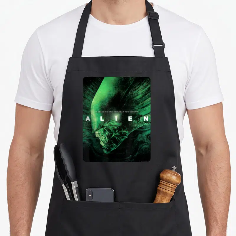 Alien movie poster apron - Green and black alien head with white text IN SPACE NO ONE CAN HEAR YOU SCREAM — shown on a black cotton canvas apron against a white background