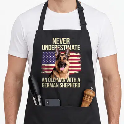 Grandpa dog mom apron - German Shepherd head over distressed USA flag with beige text reading NEVER UNDERESTIMATE — shown on a black cotton canvas apron against a white background