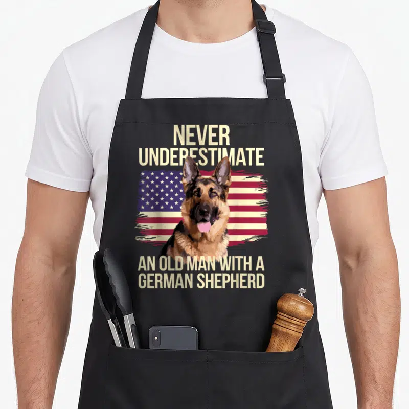 Grandpa dog mom apron - German Shepherd head over distressed USA flag with beige text reading NEVER UNDERESTIMATE — shown on a black cotton canvas apron against a white background