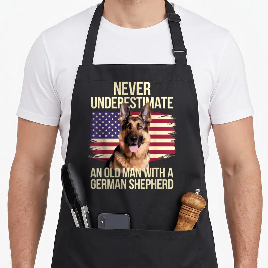 Grandpa dog mom apron - German Shepherd head over distressed USA flag with beige text reading NEVER UNDERESTIMATE — shown on a black cotton canvas apron against a white background