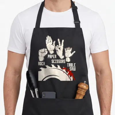 Table paper apron - Four hands labeled rock paper scissors table saw with red splatters below — shown on a black cotton canvas apron against a white background