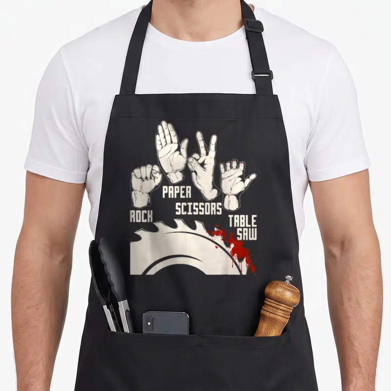 Table paper apron - Four hands labeled rock paper scissors table saw with red splatters below — shown on a black cotton canvas apron against a white background