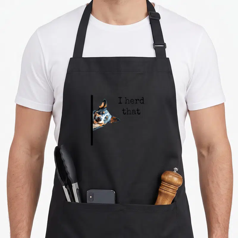 Owner dog kitchen apron - Realistic blue heeler head peeks from behind black line beside text I herd that — shown on a black cotton canvas apron against a white background