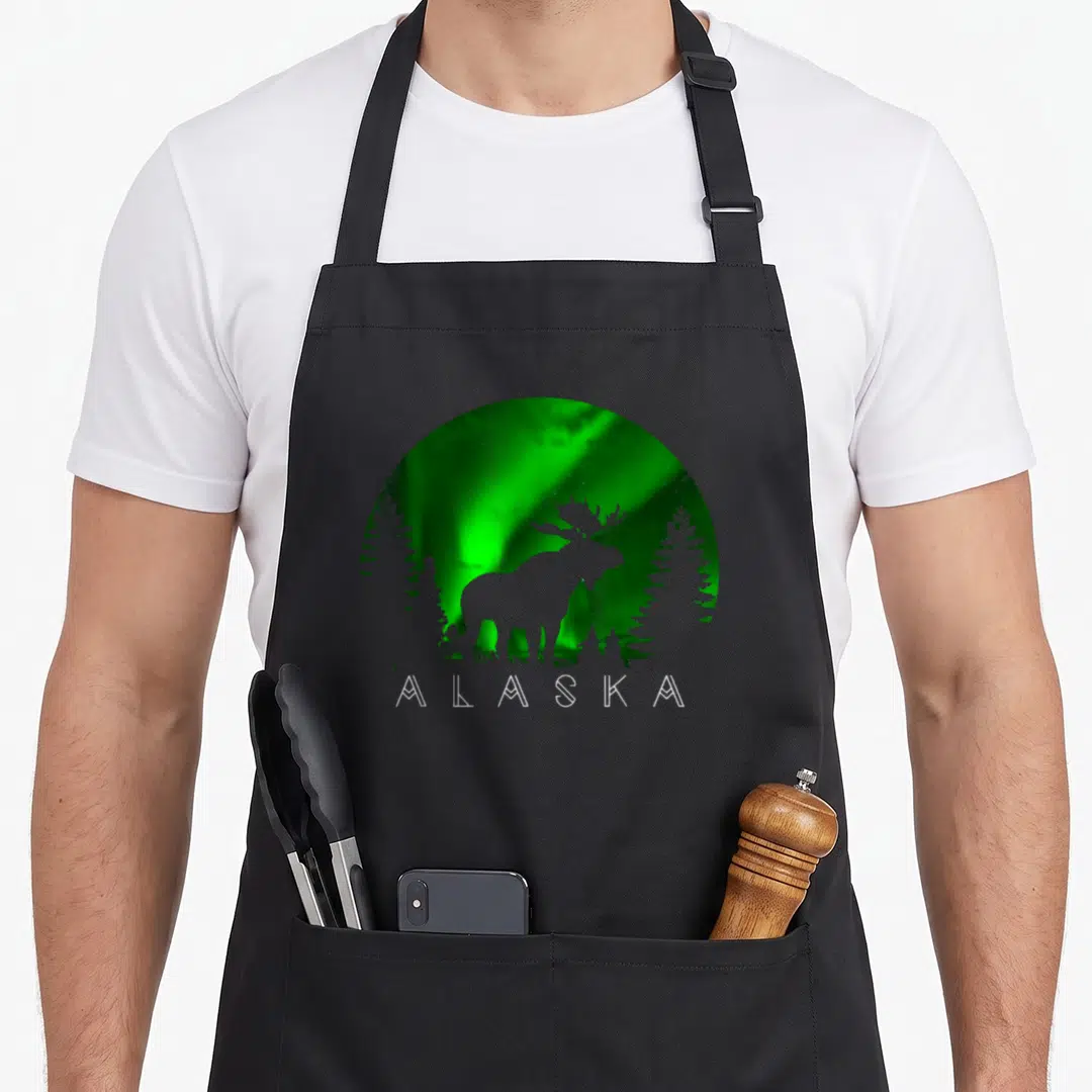 Alaska northern lights apron - Circular moose silhouette and two pine trees against green aurora with white alaska text — shown on a black cotton canvas apron against a white background