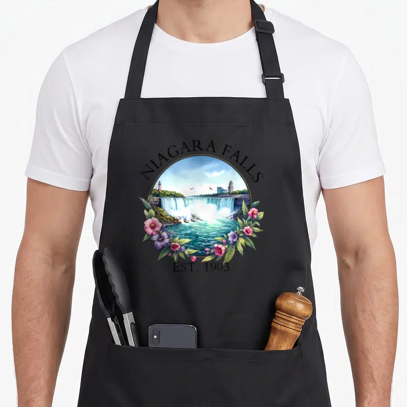 Niagara floral apron - Circular Niagara Falls landscape with pink flowers and EST 1903 text — shown on a black cotton canvas apron against a white background