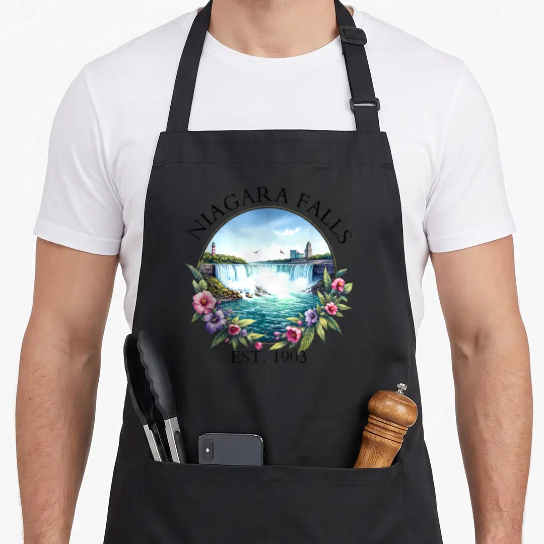 Niagara floral apron - Circular Niagara Falls landscape with pink flowers and EST 1903 text — shown on a black cotton canvas apron against a white background