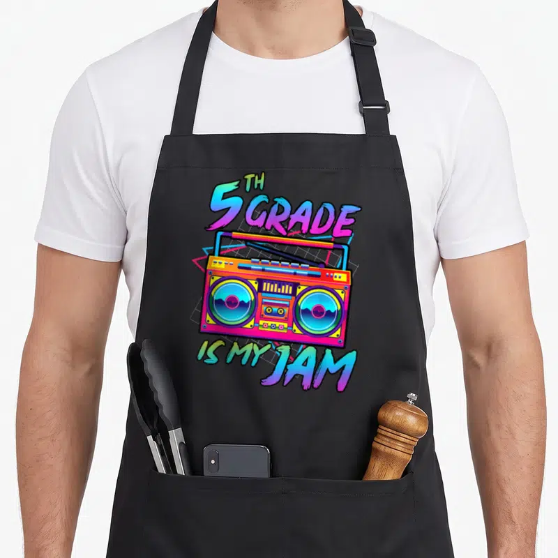 Jam art teacher apron - Retro boombox with 5th Grade Is My Jam text in pink blue orange and purple — shown on a black cotton canvas apron against a white background