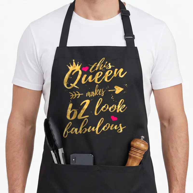 62nd queen apron - Gold script text this Queen makes 62 look fabulous with pink hearts and crown — shown on a black cotton canvas apron against a white background
