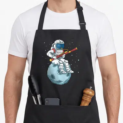 Astronaut bassoon apron - Cartoon astronaut in white suit playing red flute while sitting on a light blue moon — shown on a black cotton canvas apron against a white background