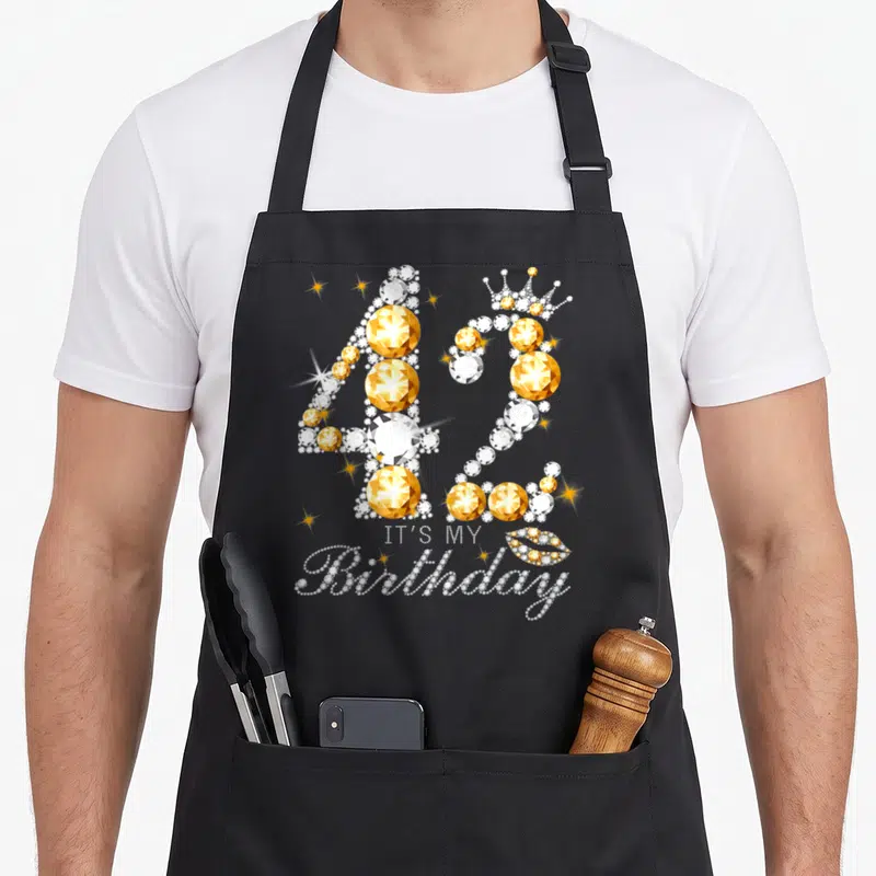 42th queen of baking apron - Diamond number 42 with crown, IT'S MY Birthday text, and jeweled lips — shown on a black cotton canvas apron against a white background