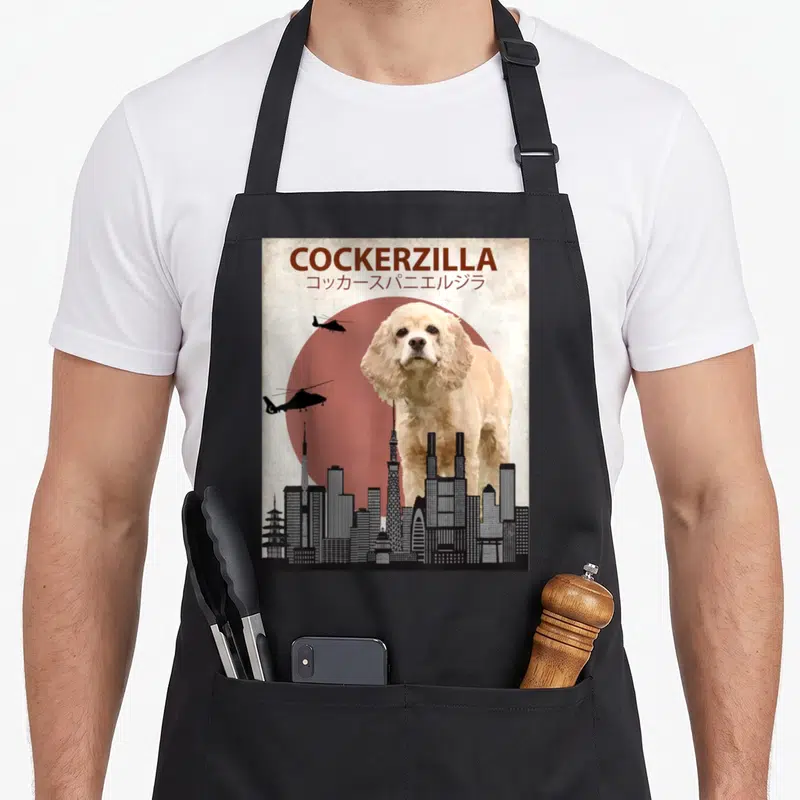 Cockerzilla cocker spaniel apron - Giant cream dog behind grey skyline with two black helicopters and brown COCKERZILLA — shown on a black cotton canvas apron against a white background