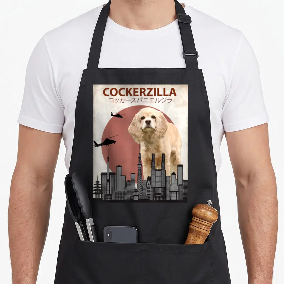 Cockerzilla cocker spaniel apron - Giant cream dog behind grey skyline with two black helicopters and brown COCKERZILLA — shown on a black cotton canvas apron against a white background