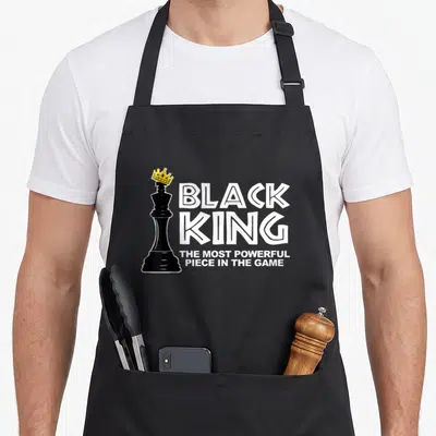 Most powerful king apron - Black chess king with yellow crown beside stacked white text reading BLACK KING THE MOST — shown on a black cotton canvas apron against a white background
