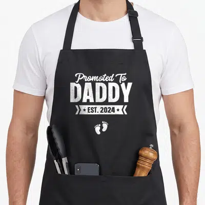 Promoted est baby daddy apron - White Promoted To Daddy EST 2024 text with baby footprints on grey background — shown on a black cotton canvas apron against a white background