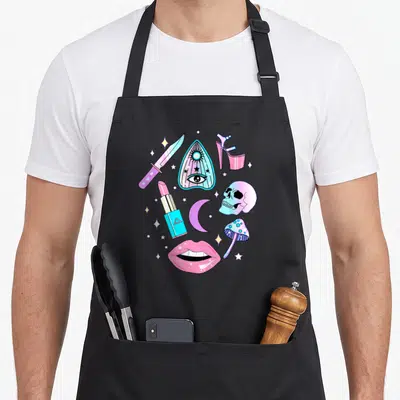Witch goth apron - Cartoon collage of pink, blue, and purple occult objects including skull, lips, and stars — shown on a black cotton canvas apron against a white background